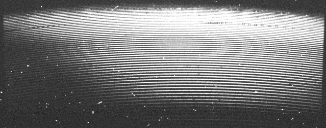 CCD-Bild eines Spektrum vom Kometen 46P/Wirtanen, aufgenommen mit dem Medium Resolution Echelle Spektrograph am Thai National Telescope. Das Spektrum sind die leicht gebogenen Linien. Die hellen und wild verteilten Punkte sind durch kosmische Strahlung, welche während der Belichtungszeit von 40 Minuten mit dem Detektor wechselwirkte, hervor gerufen. Die dunklen Stellen im Spektrum sind Absorptionslinien verschiedener Elemente oder Moleküle, das Interessanteste für die wissenschaftliche Auswertung.